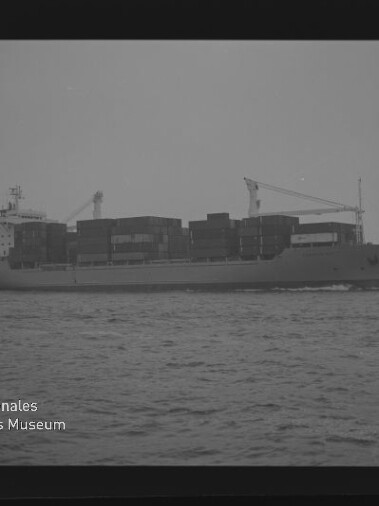 Schwarzweiß-Aufnahme eines Containerschiffs in Seitenansicht auf ruhiger See, mit gestapelten Containerblöcken und zwei Ladekränen auf dem Deck sowie sichtbarem Filmrand und Wasserzeichen des Internationalen Maritimen Museums Hamburg unten links.