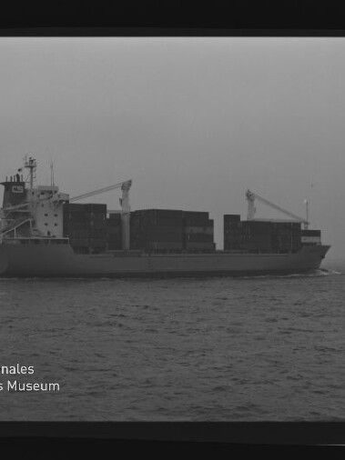 Schwarz‑weiß‑Foto eines beladenen Containerschiffs in Seitenansicht mit gestapelten Containern und zwei Ladekränen, das auf ruhiger See unter bedecktem Himmel fährt; am Bildrand Filmrand mit AGFA APX 100 und Museumswasserzeichen sichtbar.