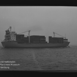 Schwarzweiß-Seitenansicht eines beladenen Containerschiffs mit aufgetürmten Containerstapeln und zwei Bordkränen, fahrend auf ruhigem Meer; Wasserzeichen des Internationalen Maritimen Museums Hamburg unten links.