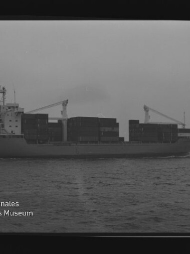 Schwarzweiß-Seitenansicht eines beladenen Containerschiffs mit aufgetürmten Containerstapeln und zwei Bordkränen, fahrend auf ruhigem Meer; Wasserzeichen des Internationalen Maritimen Museums Hamburg unten links.