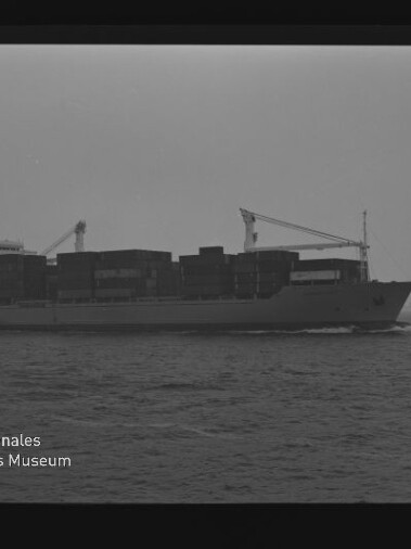 Schwarzweiß-Foto eines beladenen Containerschiffs mit aufgestapelten Containerreihen und zwei Ladekränen, das bei ruhiger See am Horizont entlangfährt; Filmrandmarkierungen und Wasserzeichen des Internationalen Maritimen Museums Hamburg sichtbar.