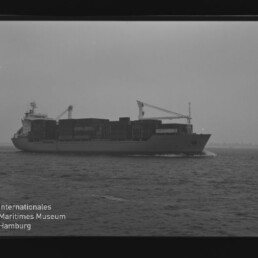 Schwarzweißfoto eines Containerschiffs mit gestapelten Containern und zwei Ladekranen, das auf ruhiger See von links nach rechts fährt; links unten sichtbar das Logo des Internationalen Maritimen Museums Hamburg und Filmrand.