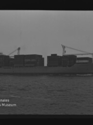 Schwarzweiß-Foto eines Containerschiffs in Seitenansicht mit aufgetürmten Containern, ruhiger See und bedecktem Himmel; unten links Logo und Schriftzug des Internationalen Maritimen Museums Hamburg.