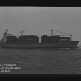 Schwarzweiß-Foto eines Containerschiffs in Seitenansicht mit aufgetürmten Containern, ruhiger See und bedecktem Himmel; unten links Logo und Schriftzug des Internationalen Maritimen Museums Hamburg.
