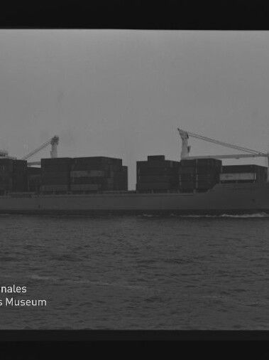 Schwarzweiß-Foto eines Containerschiffs in Seitenansicht mit aufgetürmten Containern, ruhiger See und bedecktem Himmel; unten links Logo und Schriftzug des Internationalen Maritimen Museums Hamburg.