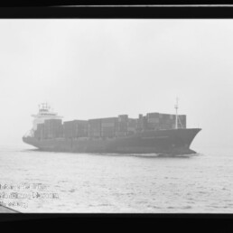 Schwarzweiß-Bild eines beladenen Containerschiffs in Seitenansicht, mit gestapelten Containerkästen auf ruhiger See unter leicht nebligem Himmel; Filmrand und Wasserzeichen des Internationalen Maritimen Museums Hamburg sind sichtbar.