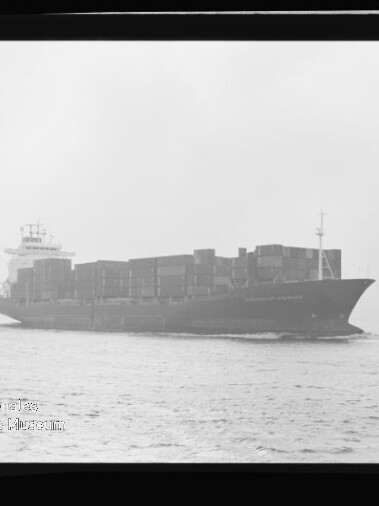 Schwarzweiß-Bild eines beladenen Containerschiffs in Seitenansicht, mit gestapelten Containerkästen auf ruhiger See unter leicht nebligem Himmel; Filmrand und Wasserzeichen des Internationalen Maritimen Museums Hamburg sind sichtbar.