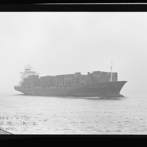 Schwarzweiß-Bild eines beladenen Containerschiffs in Seitenansicht, mit gestapelten Containerkästen auf ruhiger See unter leicht nebligem Himmel; Filmrand und Wasserzeichen des Internationalen Maritimen Museums Hamburg sind sichtbar.
