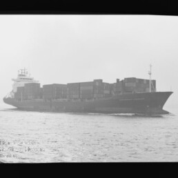 Schwarzweiß-Foto eines großen Containerschiffs auf offener See in seitlicher Ansicht, beladen mit gestapelten Containern; links unten ist ein Museumswasserzeichen sichtbar.