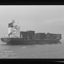 Schwarzweiß-Foto eines beladenen Containerschiffs auf See, seitlich aufgenommen mit übereinander gestapelten Containern, innerhalb eines Filmnegativ-Rahmens mit der rechten Beschriftung AGFA APX 100 und einem Wasserzeichen des Internationalen Maritimen Museums Hamburg unten links.