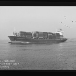 Schwarzweiß-Foto eines großen Containerschiffs mit übereinander gestapelten Containern, das über ruhige See fährt und eine kleine Bugwelle zieht; Aufnahme zeigt Filmrand, leichte Beschädigungen im Himmel und das Wasserzeichen des Internationalen Maritimen Museums Hamburg unten links.