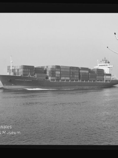 Schwarzweiß-Foto eines großen Containerschiffs mit übereinander gestapelten Containern, das über ruhige See fährt und eine kleine Bugwelle zieht; Aufnahme zeigt Filmrand, leichte Beschädigungen im Himmel und das Wasserzeichen des Internationalen Maritimen Museums Hamburg unten links.