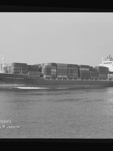 Schwarzweiß-Aufnahme eines großen Containerschiffs, beladen mit gestapelten Containerreihen, das bei ruhiger See mit sichtbarer Bugwelle dahinfährt; schwarzer Filmrand und Museumslogo links unten erkennbar.
