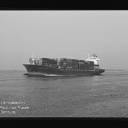 Schwarzweiß-Foto eines großen Containerschiffs mit gestapelten Containern, das auf ruhiger See von links nach rechts fährt; Filmrand und Museum-Logo unten links sichtbar.