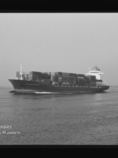 Schwarzweiß-Foto eines großen Containerschiffs mit gestapelten Containern, das auf ruhiger See von links nach rechts fährt; Filmrand und Museum-Logo unten links sichtbar.
