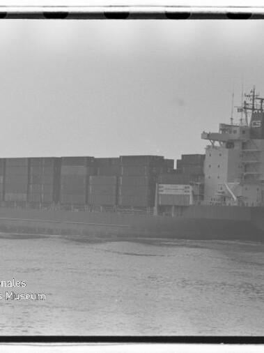 Schwarzweißaufnahme eines großen Containerschiffs mit übereinander gestapelten Ladecontainern, das bei ruhiger See von rechts nach links fährt; sichtbarer Filmrand und Museumswasserzeichen unten links.