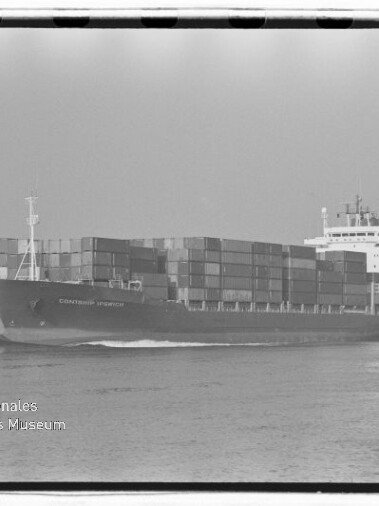 Schwarzweiß-Aufnahme eines beladenen Containerschiffs, das mit hohen Stapeln von Frachtcontainern auf ruhiger See fährt; am unteren linken Bildrand ein Wasserzeichen des Internationalen Maritimen Museums Hamburg.