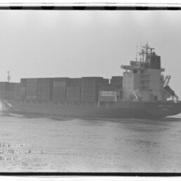 Schwarzweiß-Foto eines großen Containerschiffs mit übereinander gestapelten Frachtcontainern und einem hohen Deckshaus am Heck, das ruhig durch leicht nebliges Wasser fährt, links unten sichtbarer Stempel des Internationalen Maritimen Museums Hamburg