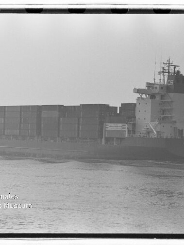 Schwarzweiß-Foto eines großen Containerschiffs mit übereinander gestapelten Frachtcontainern und einem hohen Deckshaus am Heck, das ruhig durch leicht nebliges Wasser fährt, links unten sichtbarer Stempel des Internationalen Maritimen Museums Hamburg