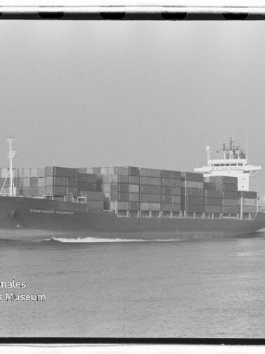 Schwarzweißfoto eines großen Containerschiffs, dicht mit übereinander gestapelten Frachtcontainern beladen, das bei ruhiger See entlangfährt; unten links sichtbar das Wasserzeichen des Internationalen Maritimen Museums Hamburg.