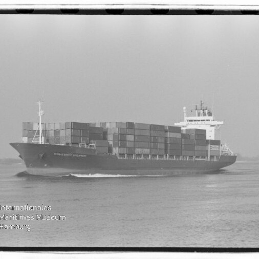 Schwarzweißfoto eines großen Containerschiffs, dicht mit übereinander gestapelten Frachtcontainern beladen, das bei ruhiger See entlangfährt; unten links sichtbar das Wasserzeichen des Internationalen Maritimen Museums Hamburg.