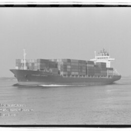 Schwarzweißaufnahme eines großen Containerschiffs, beladen mit mehrfach übereinander gestapelten Ladecontainern und mit sichtbarer Brücke, das auf ruhigem Meer fährt; links unten Logo des Internationalen Maritimen Museums Hamburg.