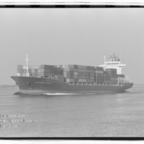 Schwarzweißaufnahme eines großen Containerschiffs, beladen mit mehrfach übereinander gestapelten Ladecontainern und mit sichtbarer Brücke, das auf ruhigem Meer fährt; links unten Logo des Internationalen Maritimen Museums Hamburg.