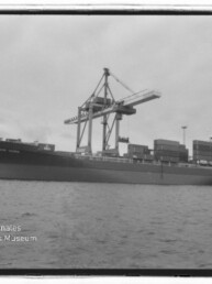 Schwarzweißaufnahme eines großen Containerschiffs am Kai mit gestapelten Containern und hohen Verladekränen, im Vordergrund Wasser und Wasserzeichen des Internationalen Maritimen Museums Hamburg.