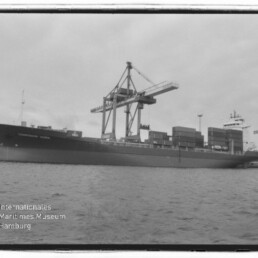 Schwarzweißaufnahme eines großen Containerschiffs am Kai mit gestapelten Containern und hohen Verladekränen, im Vordergrund Wasser und Wasserzeichen des Internationalen Maritimen Museums Hamburg.