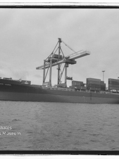 Schwarzweißaufnahme eines großen Containerschiffs am Kai mit gestapelten Containern und hohen Verladekränen, im Vordergrund Wasser und Wasserzeichen des Internationalen Maritimen Museums Hamburg.