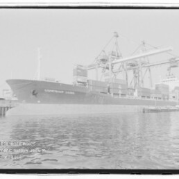 Containerschiff CONTSHIP JORK liegt am Kai eines Hafens unter großen Brückenkränen, gestapelte Container an Deck und ruhiges Wasser im Vordergrund; Stempel Internationales Maritimes Museum Hamburg unten links.