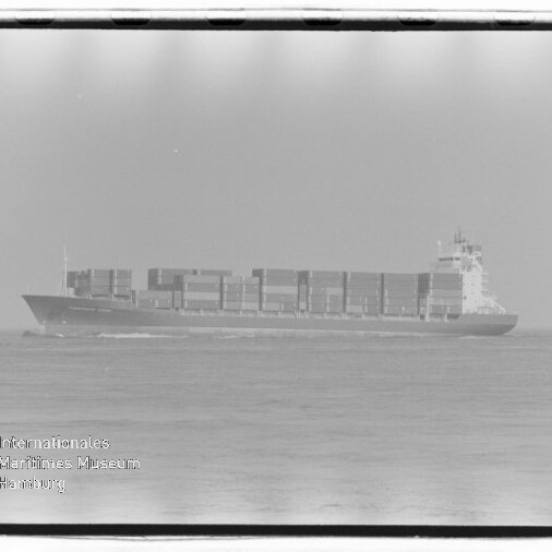 Schwarzweiß-Aufnahme eines Containerschiffs in Seitenansicht mit mehreren übereinander gestapelten Containerreihen, das ruhig auf dem Meer fährt; unten links sichtbar das Wasserzeichen des Internationalen Maritimen Museums Hamburg und ein Filmrandrahmen.