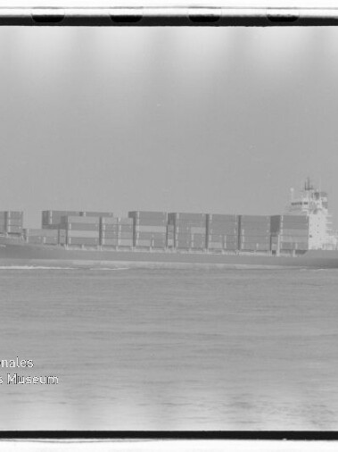 Schwarzweiß-Seitenansicht eines beladenen Containerschiffs auf ruhiger See mit gestapelten Containern und sichtbarer Bugwelle; unten links Wasserzeichen „Internationales Maritimes Museum Hamburg“.