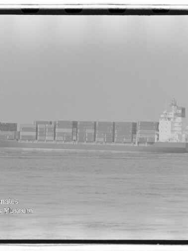Schwarzweißaufnahme eines Containerschiffs auf ruhiger See mit übereinander gestapelten Containern und einem Aufbau am Heck, vor hellem Himmel; unten links sichtbar das Wasserzeichen 