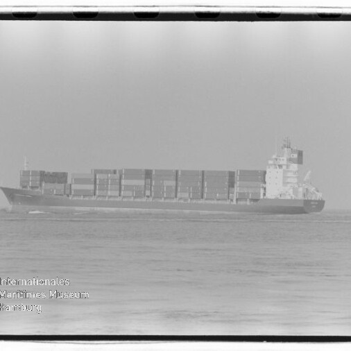 Schwarzweißaufnahme eines Containerschiffs auf ruhiger See mit übereinander gestapelten Containern und einem Aufbau am Heck, vor hellem Himmel; unten links sichtbar das Wasserzeichen 