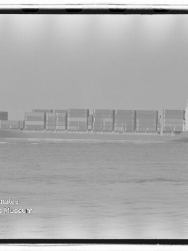Schwarzweiß-Foto eines seitenansichtigen Containerschiffs auf ruhiger See mit gestapelten Containern, sichtbarem Filmrand und Museumswasserzeichen unten links.