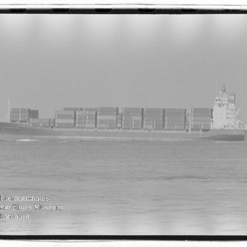 Schwarzweiß-Foto eines seitenansichtigen Containerschiffs auf ruhiger See mit gestapelten Containern, sichtbarem Filmrand und Museumswasserzeichen unten links.
