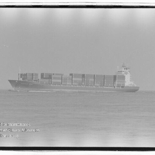 Containerschiff mit aufgestapelten Ladecontainern fährt auf ruhiger See, im Vordergrund rechts eine Möwe und unten links ein Wasserzeichen des Internationalen Maritimen Museums Hamburg.
