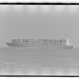 Schwarzweiß-Foto eines großen Containerschiffs auf ruhiger See, mittig im Bild mit mehreren Reihen gestapelter Container, sichtbarem Horizont und Filmnegativrand; unten links Museumswasserzeichen 'Internationales Maritimes Museum Hamburg'.