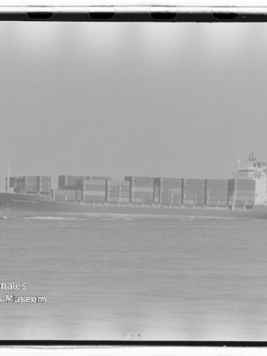 Schwarzweiß-Foto eines großen Containerschiffs auf ruhiger See, mittig im Bild mit mehreren Reihen gestapelter Container, sichtbarem Horizont und Filmnegativrand; unten links Museumswasserzeichen 'Internationales Maritimes Museum Hamburg'.