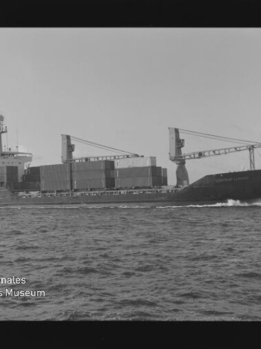 Schwarzweißaufnahme eines Containerschiffs mit gestapelten Containern und zwei Ladekränen, das mit sichtbarer Bugwelle übers Meer fährt; unten links Wasserzeichen des Internationalen Maritimen Museums Hamburg.