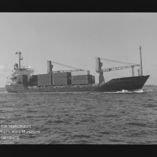 Schwarzweißaufnahme eines Containerschiffs mit gestapelten Containern und zwei Ladekränen, das mit sichtbarer Bugwelle übers Meer fährt; unten links Wasserzeichen des Internationalen Maritimen Museums Hamburg.
