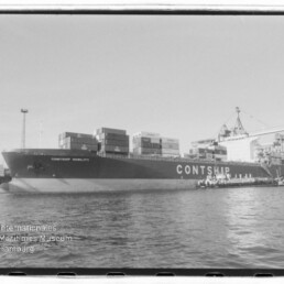 Großes Containerschiff mit der Aufschrift CONTSHIP, beladen mit Containern und am Kai liegend, davor ruhiges Wasser und im Hintergrund Hafenkräne; Wasserzeichen des Internationalen Maritimen Museums Hamburg in der unteren linken Ecke.