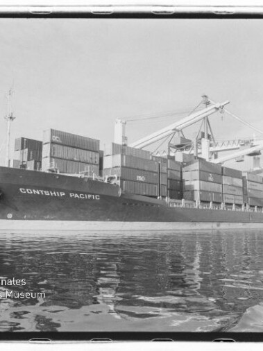 Schwarzweißaufnahme des Containerschiffs 'CONTSHIP PACIFIC' am Kai, beladen mit gestapelten Containern, umgeben von Hafenkranen und mit Reflexion im Wasser; links unten das Logo des Internationalen Maritimen Museums Hamburg.