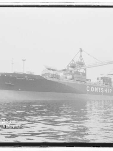 Schwarzweißfoto eines großen Containerschiffs mit der auffälligen Schrift „CONTSHIP“ am Kai, umgeben von Hafenkränen und beladenen Containern, mit ruhiger Wasseroberfläche im Vordergrund und einem Museumswasserzeichen unten links.