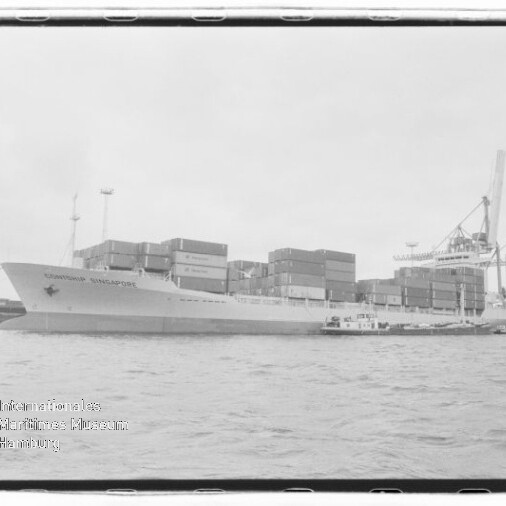 Großes Containerschiff mit gestapelten Ladecontainern liegt im Hafen, rechts Kräne im Hintergrund und ein kleines Begleitschiff längsseits, Wasser im Vordergrund und ein Wasserzeichen des Internationalen Maritimen Museums Hamburg unten links.
