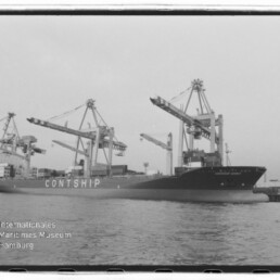 Schwarzweißaufnahme eines großen Containerschiffs mit der Aufschrift CONTSHIP, das am Hafen unter mehreren Containerkränen liegt; Wasser im Vordergrund und das Logo des Internationalen Maritimen Museums Hamburg unten links.