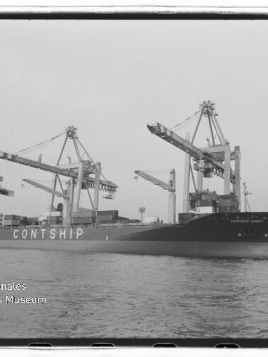 Schwarzweißaufnahme eines großen Containerschiffs mit der Aufschrift CONTSHIP, das am Hafen unter mehreren Containerkränen liegt; Wasser im Vordergrund und das Logo des Internationalen Maritimen Museums Hamburg unten links.