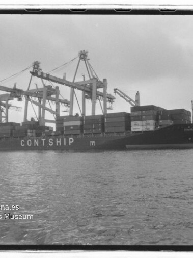 Schwarzweißaufnahme eines beladenen Containerschiffs mit dem Schriftzug CONTSHIP, das am Kai von mehreren Hafenkranen be- oder entladen wird; im Vordergrund Wasser und links unten das Logo des Internationalen Maritimen Museums Hamburg.