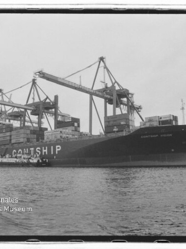 Schwarzweißaufnahme eines großen Containerschiffs mit der Aufschrift CONTSHIP, das am Hafen von mehreren Containerkränen be- oder entladen wird, vor ruhigem Wasser.